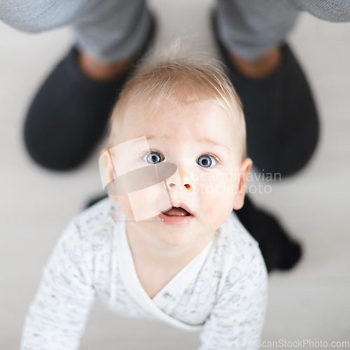 Image of Top down view of cheerful baby boy infant taking first steps holding to father's sweatpants at home. Cute baby boy learning to walk