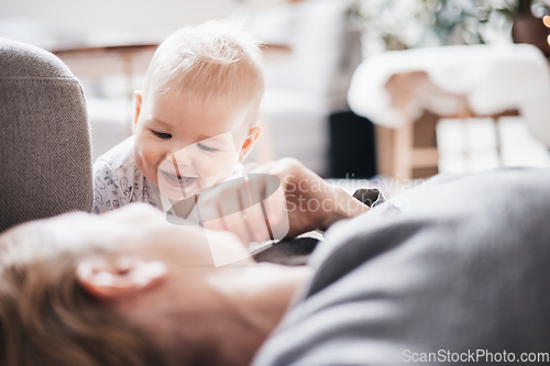 Image of Happy family moments. Mother lying comfortably on children's mat playing with her baby boy watching and suppervising his first steps. Positive human emotions, feelings, joy.