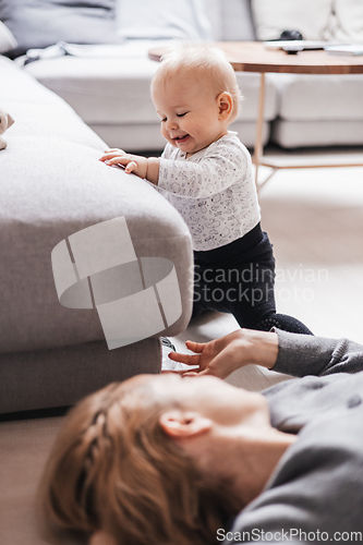 Image of Happy family moments. Mother lying comfortably on children's mat playing with her baby boy watching and suppervising his first steps. Positive human emotions, feelings, joy.