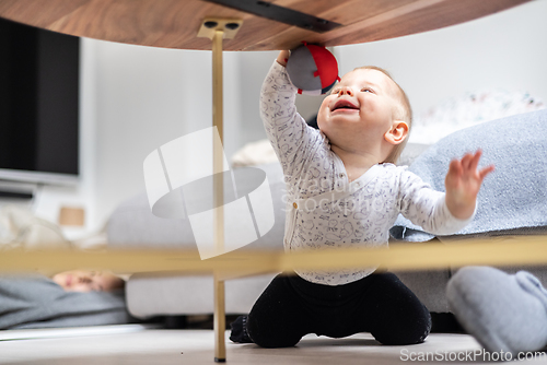 Image of Cute infant baby boy playing with hanging ball, crawling and standing up by living room table at home. Baby activity and play center for early infant development. Baby playing at home.