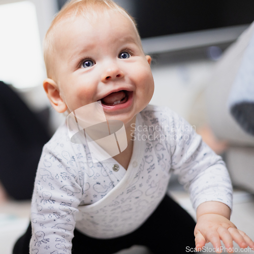 Image of Cute infant baby boy playing, crawling and standing up by living room sofa at home. Baby playing at home