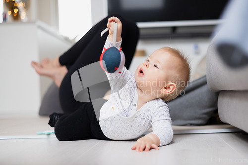 Image of Happy family moments. Mother lying comfortably on children's mat watching and suppervising her baby boy playinghis in living room. Positive human emotions, feelings, joy