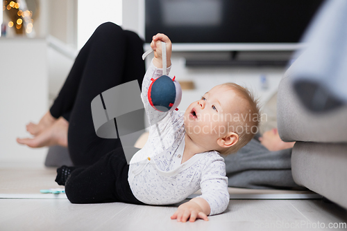 Image of Happy family moments. Mother lying comfortably on children's mat watching and suppervising her baby boy playinghis in living room. Positive human emotions, feelings, joy