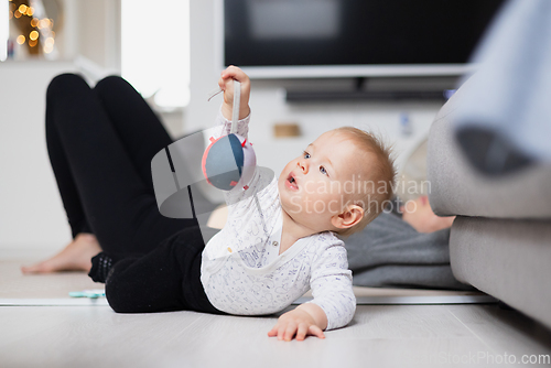 Image of Happy family moments. Mother lying comfortably on children's mat watching and suppervising her baby boy playinghis in living room. Positive human emotions, feelings, joy