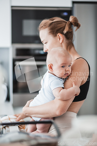 Image of Woman wiping kitchen sink with a cloth after finishing washing the dishes while holding four months old baby boy in her hands