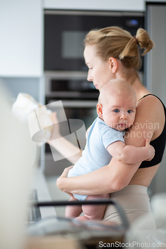 Image of Woman wiping kitchen sink with a cloth after finishing washing the dishes while holding four months old baby boy in her hands