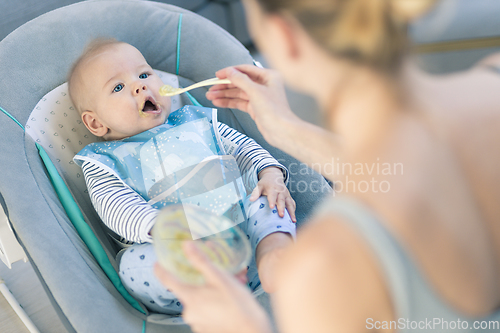 Image of Mother spoon feeding her baby boy infant child in baby chair with fruit puree. Baby solid food introduction concept.