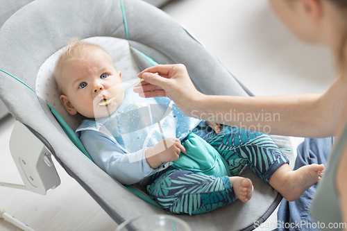 Image of Mother spoon feeding her baby boy infant child in baby chair with fruit puree. Baby solid food introduction concept.