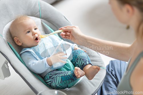 Image of Mother spoon feeding her baby boy infant child in baby chair with fruit puree. Baby solid food introduction concept.