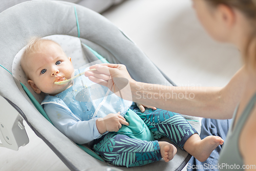 Image of Mother spoon feeding her baby boy infant child in baby chair with fruit puree. Baby solid food introduction concept.