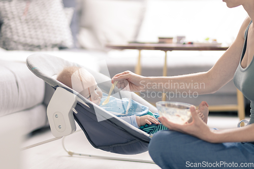 Image of Mother spoon feeding her baby boy infant child in baby chair with fruit puree. Baby solid food introduction concept.