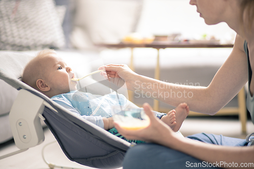 Image of Mother spoon feeding her baby boy infant child in baby chair with fruit puree. Baby solid food introduction concept.