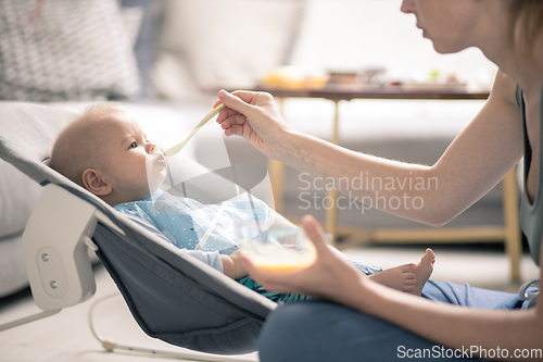 Image of Mother spoon feeding her baby boy infant child in baby chair with fruit puree. Baby solid food introduction concept.