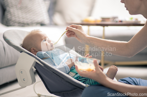 Image of Mother spoon feeding her baby boy infant child in baby chair with fruit puree. Baby solid food introduction concept.