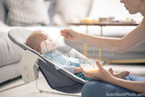 Image of Mother spoon feeding her baby boy infant child in baby chair with fruit puree. Baby solid food introduction concept.