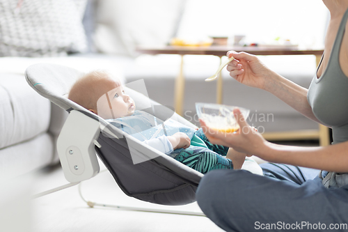 Image of Mother spoon feeding her baby boy infant child in baby chair with fruit puree. Baby solid food introduction concept.