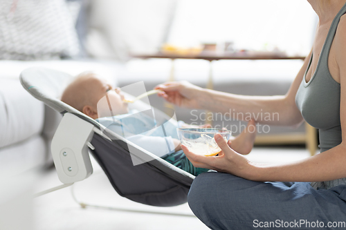 Image of Mother spoon feeding her baby boy infant child in baby chair with fruit puree. Baby solid food introduction concept.