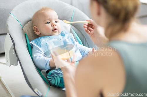 Image of Mother spoon feeding her baby boy infant child in baby chair with fruit puree. Baby solid food introduction concept.