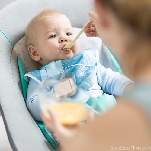 Image of Mother spoon feeding her baby boy infant child in baby chair with fruit puree. Baby solid food introduction concept.