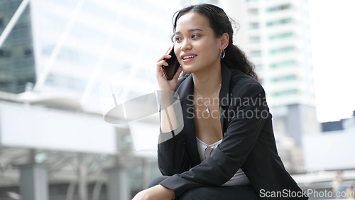 Image of A young Asian businesswoman in formal attire sits smiling and using a smartphone