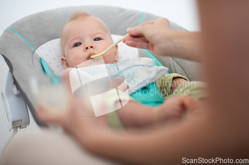 Image of Mother spoon feeding her baby boy infant child in baby chair with fruit puree. Baby solid food introduction concept.