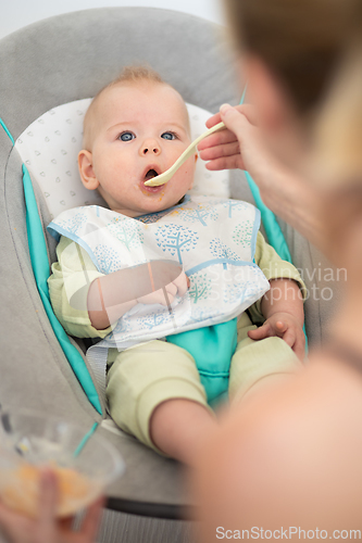 Image of Mother spoon feeding her baby boy infant child in baby chair with fruit puree. Baby solid food introduction concept.