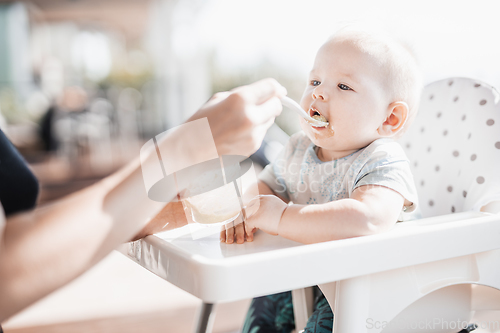 Image of Mother spoon feeding her baby boy child in baby chair with fruit puree on a porch on summer vacations. Baby solid food introduction concept.