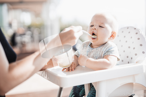 Image of Mother spoon feeding her baby boy child in baby chair with fruit puree on a porch on summer vacations. Baby solid food introduction concept.