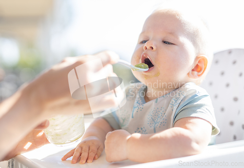 Image of Mother spoon feeding her baby boy child in baby chair with fruit puree on a porch on summer vacations. Baby solid food introduction concept.