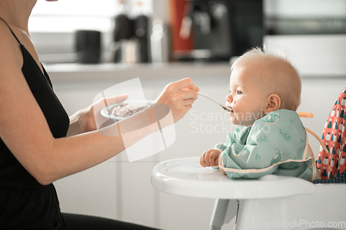 Image of Mother spoon feeding her baby boy child in baby chair with fruit puree in kitchen at home. Baby solid food introduction concept.