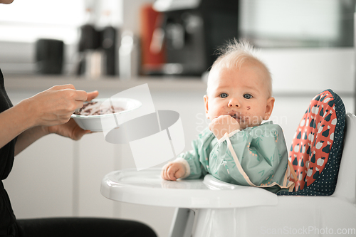 Image of Mother spoon feeding her baby boy child in baby chair with fruit puree in kitchen at home. Baby solid food introduction concept.