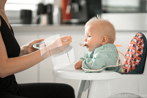 Image of Mother spoon feeding her baby boy child in baby chair with fruit puree in kitchen at home. Baby solid food introduction concept.