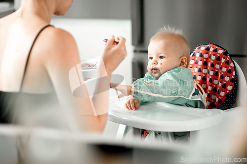 Image of Mother spoon feeding her baby boy child in baby chair with fruit puree in kitchen at home. Baby solid food introduction concept.