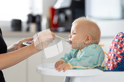 Image of Mother spoon feeding her baby boy child in baby chair with fruit puree in kitchen at home. Baby solid food introduction concept.