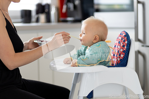 Image of Mother spoon feeding her baby boy child in baby chair with fruit puree in kitchen at home. Baby solid food introduction concept.