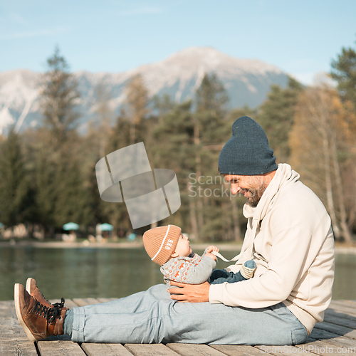 Image of Happy family. Father playing with her baby boy infant oudoors on sunny autumn day. Portrait of dad and little son on wooden platform by lake. Positive human emotions, feelings, joy.