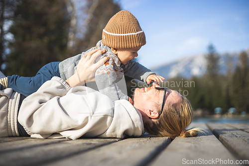 Image of Happy family. Young mother playing with her baby boy infant oudoors on sunny autumn day. Portrait of mom and little son on wooden platform by lake. Positive human emotions, feelings, joy.