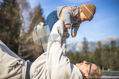 Image of Happy family. Young mother playing with her baby boy infant oudoors on sunny autumn day. Portrait of mom and little son on wooden platform by lake. Positive human emotions, feelings, joy.