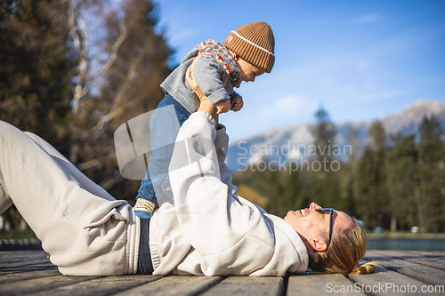 Image of Happy family. Young mother playing with her baby boy infant oudoors on sunny autumn day. Portrait of mom and little son on wooden platform by lake. Positive human emotions, feelings, joy.