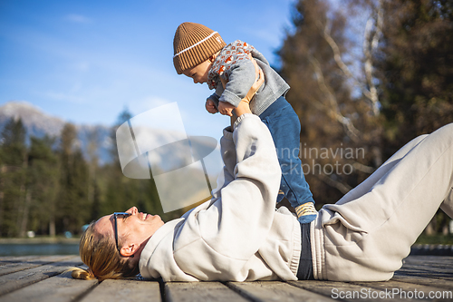 Image of Happy family. Young mother playing with her baby boy infant oudoors on sunny autumn day. Portrait of mom and little son on wooden platform by lake. Positive human emotions, feelings, joy.