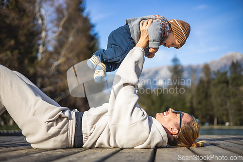 Image of Happy family. Young mother playing with her baby boy infant oudoors on sunny autumn day. Portrait of mom and little son on wooden platform by lake. Positive human emotions, feelings, joy.