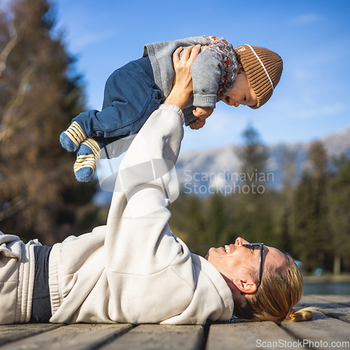 Image of Happy family. Young mother playing with her baby boy infant oudoors on sunny autumn day. Portrait of mom and little son on wooden platform by lake. Positive human emotions, feelings, joy.