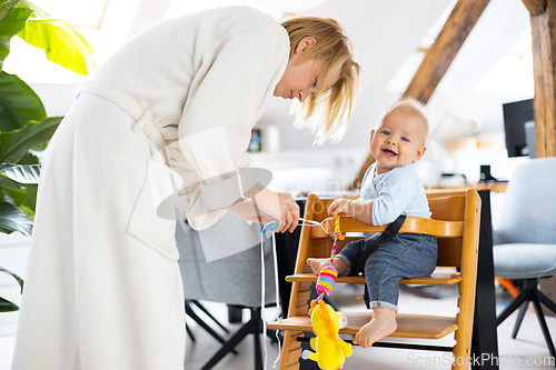 Image of Happy infant sitting and playing with his toy in traditional scandinavian designer wooden high chair in modern bright atic home suppervised by his mother.