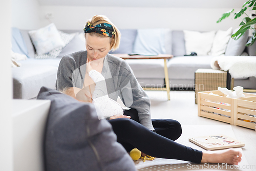 Image of Young woman breastfeeding her infant baby boy casualy sitting on child's playing mat on living room floor at home.