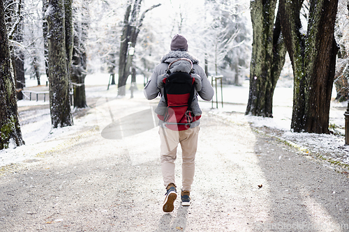 Image of Rear view of sporty father carrying his infant son wearing winter jumpsuit and cap in backpack carrier walking in city park in winter