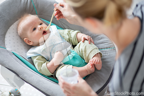 Image of Mother spoon feeding her baby boy infant child in baby chair with fruit puree. Baby solid food introduction concept.