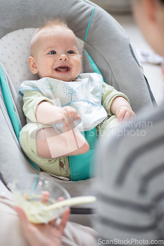 Image of Mother spoon feeding her baby boy infant child in baby chair with fruit puree. Baby solid food introduction concept.