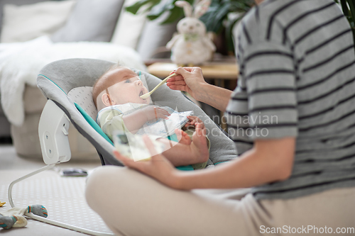 Image of Mother spoon feeding her baby boy infant child in baby chair with fruit puree. Baby solid food introduction concept.