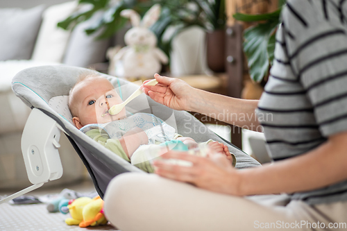 Image of Mother spoon feeding her baby boy infant child in baby chair with fruit puree. Baby solid food introduction concept.