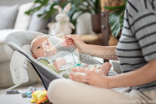 Image of Mother spoon feeding her baby boy infant child in baby chair with fruit puree. Baby solid food introduction concept.